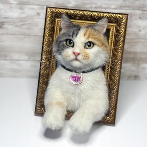 May include: A needle-felted calico cat sculpture with white, gray, and orange fur, positioned inside an ornate gold-colored rectangular frame. The cat wears a black collar with a clear, cat-shaped pendant featuring the name Lotti in pink text.