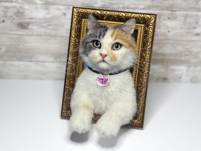 May include: A needle-felted calico cat sculpture with white, gray, and orange fur, positioned inside an ornate gold-colored rectangular frame. The cat wears a black collar with a clear, cat-shaped pendant featuring the name Lotti in pink text.