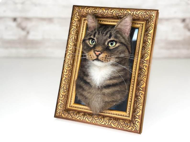 May include: A three-dimensional needle felted sculpture of a brown tabby cat with a white chest, mounted inside an ornate, rectangular, antique-style gold frame with intricate scrollwork patterns along the border and a dark inner background.