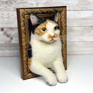 May include: A needle-felted calico cat sculpture emerging from a rectangular, ornate, antique-style gold frame. The cat has white, black, and orange fur, green eyes, and a pink nose, with its front paws resting over the bottom edge of the frame.