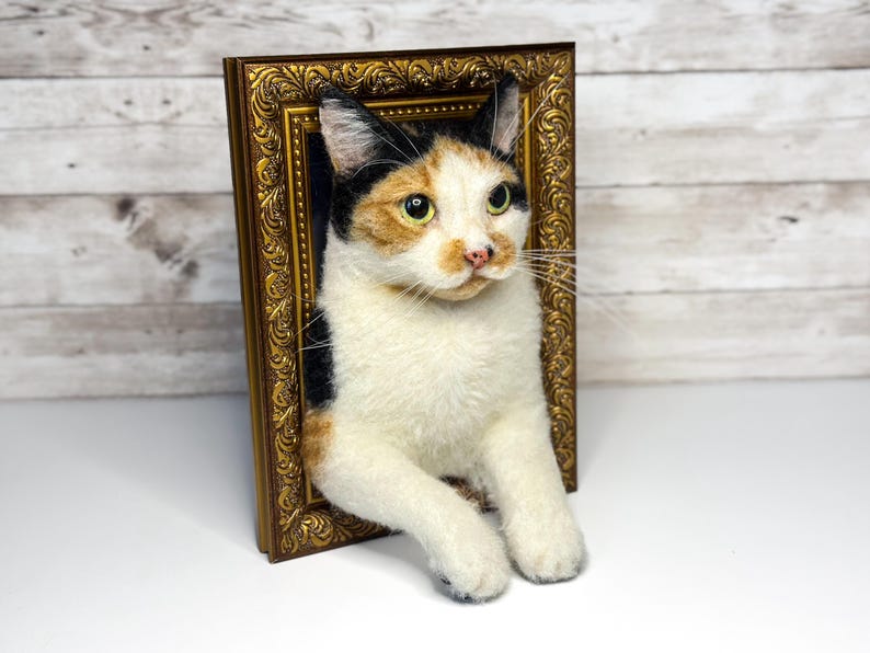May include: A needle-felted calico cat sculpture emerging from a rectangular, ornate, antique-style gold frame. The cat has white, black, and orange fur, green eyes, and a pink nose, with its front paws resting over the bottom edge of the frame.