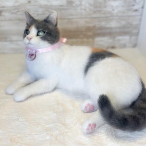 May include: A handmade cat figurine in a relaxed pose. The cat has a white body with gray and orange markings. It wears a pink collar with a charm. The cat's paws are pink, and its tail is gray. The cat is lying on a soft, white surface.