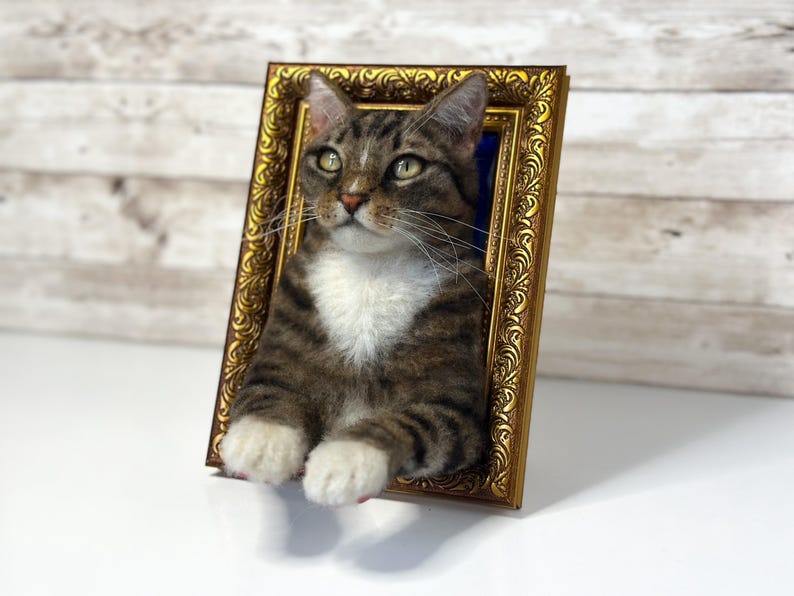 Un gato atigrado marrón de fieltro de aguja tridimensional con pecho y patas blancas, que emerge de un marco de fotos rectangular, ornamentado y de color dorado con intrincados relieves en el borde.