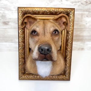 May include: A three-dimensional needle felted sculpture of a tan dog head with a white chest patch, brown eyes, and a black nose, mounted inside a rectangular, ornate, metallic gold-coloured picture frame with intricate scrollwork detailing.