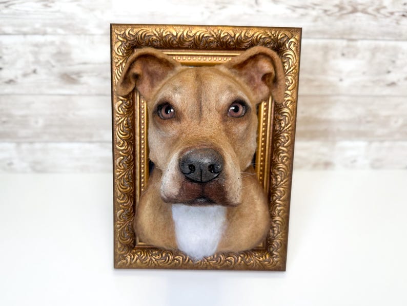 May include: A three-dimensional needle felted sculpture of a tan dog head with a white chest patch, brown eyes, and a black nose, mounted inside a rectangular, ornate, metallic gold-coloured picture frame with intricate scrollwork detailing.