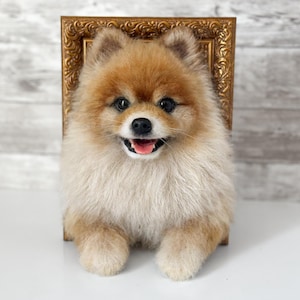 May include: A needle felted Pomeranian dog sculpture with tan and cream fur, black eyes, and a black nose, positioned in front of a small, ornate, rectangular gold-coloured picture frame with intricate carved detailing along the edges.