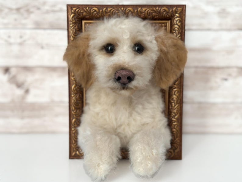 May include: A needle-felted sculpture of a cream-coloured dog with light brown ears and a pink nose, positioned as if emerging from a rectangular, ornate, antique-style bronze-coloured picture frame with intricate scrollwork detailing.