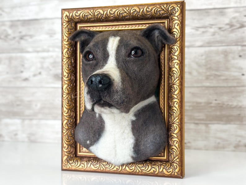 May include: A three-dimensional needle felted sculpture of a grey and white pit bull dog head mounted inside an ornate, rectangular gold-coloured frame. The dog has dark eyes, a black nose, and a white stripe running from its forehead to its chest.