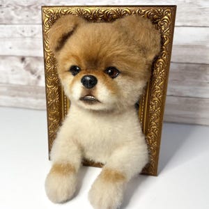 May include: A needle-felted sculpture of a small dog with tan and white fur, featuring dark eyes and a black nose, emerging from a rectangular, ornate, metallic gold-coloured picture frame that creates a three-dimensional wall-mounted effect.