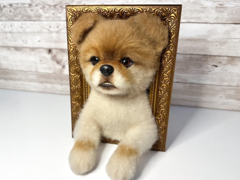 May include: A needle-felted sculpture of a small dog with tan and white fur, featuring dark eyes and a black nose, emerging from a rectangular, ornate, metallic gold-coloured picture frame that creates a three-dimensional wall-mounted effect.