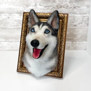 May include: A needle-felted husky dog head sculpture mounted inside an ornate, rectangular, metallic gold-coloured frame. The husky features grey and white fur, bright blue eyes, a black nose, and an open mouth showing a pink tongue.