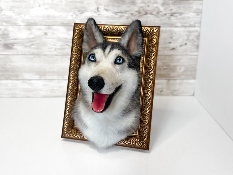May include: A needle-felted husky dog head sculpture mounted inside an ornate, rectangular, metallic gold-coloured frame. The husky features grey and white fur, bright blue eyes, a black nose, and an open mouth showing a pink tongue.