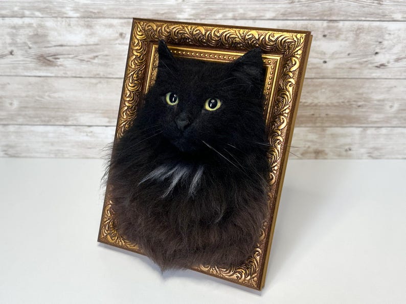 May include: A three-dimensional wall art piece featuring a realistic, fluffy black cat head with bright yellow-green eyes and a small patch of white fur on the chest, mounted inside an ornate, rectangular, metallic gold-colored picture frame.