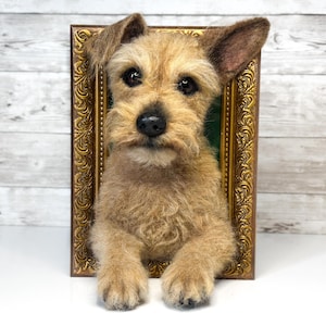 May include: A needle-felted tan terrier dog sculpture emerging from a rectangular, ornate gold-coloured picture frame. The dog has a textured, shaggy coat, dark eyes, a black nose, and one ear standing upright while the other is folded down.