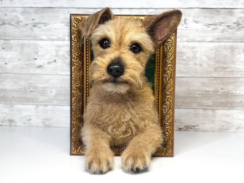 May include: A needle-felted tan terrier dog sculpture emerging from a rectangular, ornate gold-coloured picture frame. The dog has a textured, shaggy coat, dark eyes, a black nose, and one ear standing upright while the other is folded down.