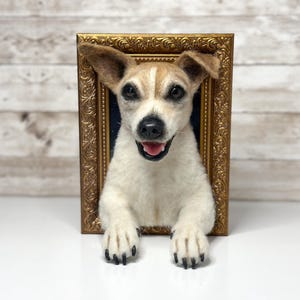 May include: A needle-felted Jack Russell Terrier sculpture emerging from a rectangular, ornate gold-coloured frame. The dog has white fur with tan markings on its head and ears, dark eyes, a black nose, and an open mouth showing a pink tongue.