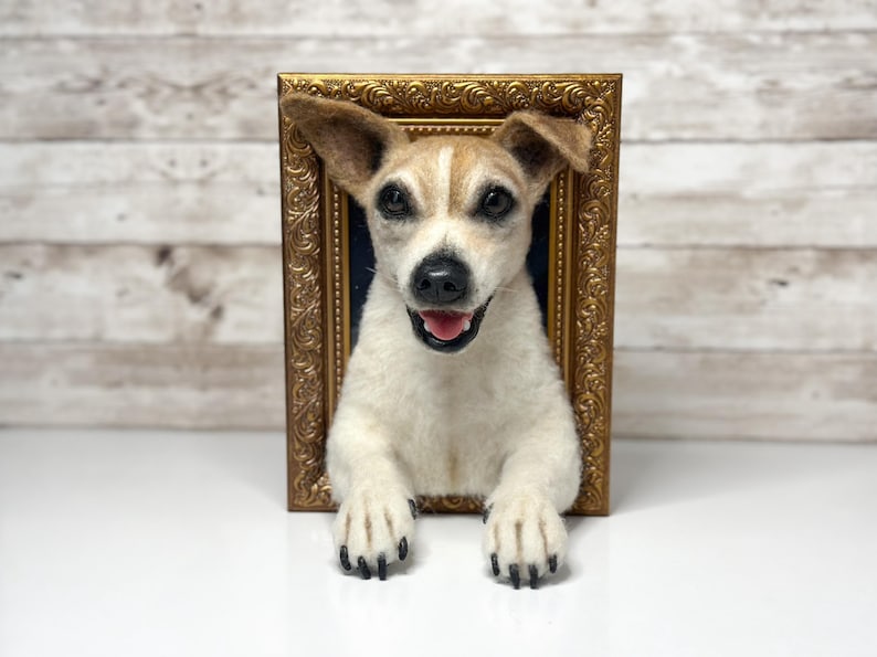 May include: A needle-felted Jack Russell Terrier sculpture emerging from a rectangular, ornate gold-coloured frame. The dog has white fur with tan markings on its head and ears, dark eyes, a black nose, and an open mouth showing a pink tongue.