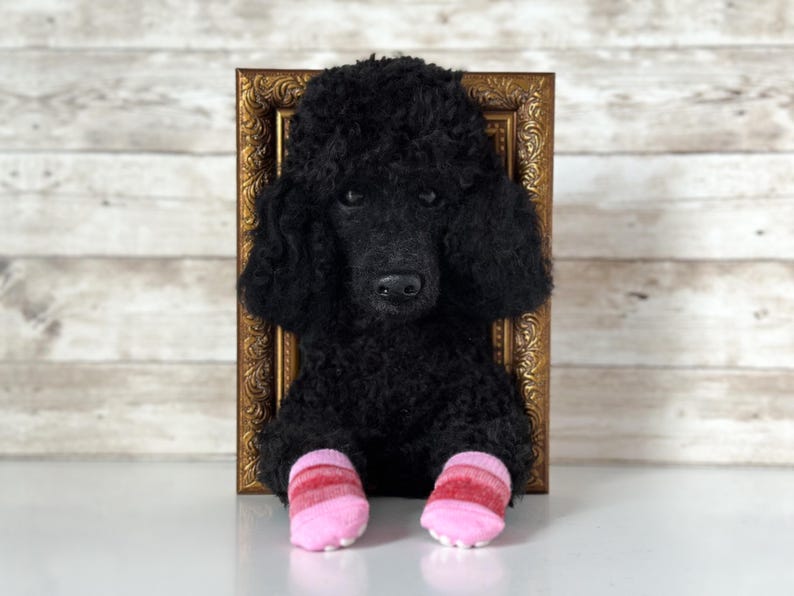 May include: A black poodle plush toy with curly fur positioned in front of an ornate gold rectangular frame. The plush toy has dark eyes and is wearing two small, striped pink and red socks on its front paws, with light pink toe grips visible.