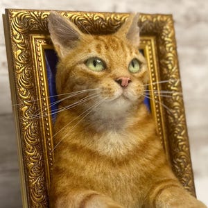 May include: A needle-felted orange tabby cat sculpture with green eyes and a pink nose, positioned within an ornate, rectangular gold-colored frame. The cat has detailed fur texture, long white whiskers, and is set against a dark blue background.