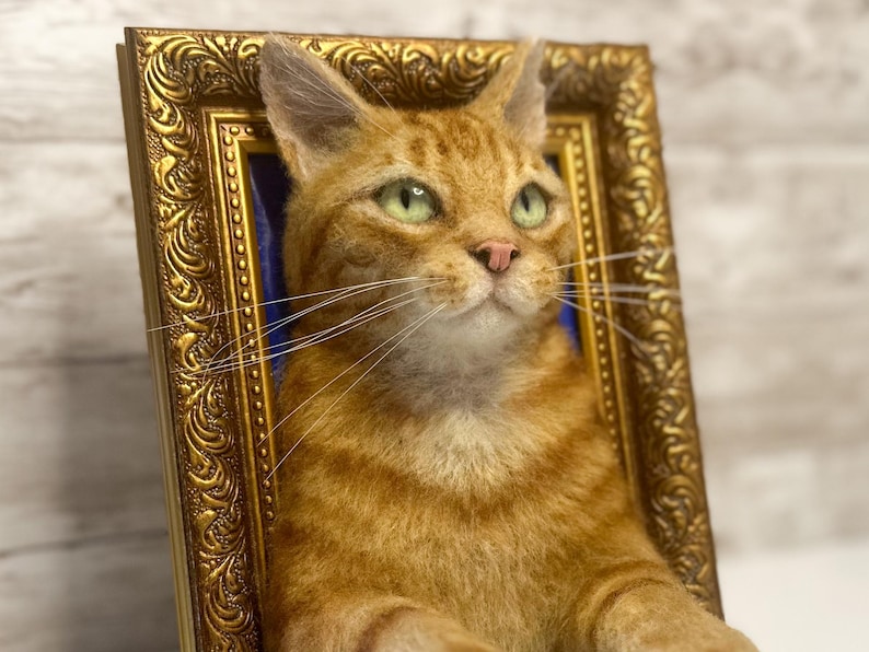 May include: A needle-felted orange tabby cat sculpture with green eyes and a pink nose, positioned within an ornate, rectangular gold-colored frame. The cat has detailed fur texture, long white whiskers, and is set against a dark blue background.