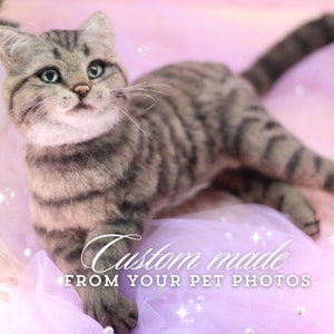 Puede incluir: Escultura de gato realista y personalizada en tonos grises y marrones, con ojos verdes y bigotes largos. El gato está tumbado sobre una superficie rosa y morada. El texto de la imagen dice "Custom made from your pet photos."