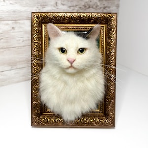May include: A three-dimensional needle felted white cat head sculpture mounted inside an ornate, rectangular, metallic gold-colored picture frame. The cat has long white fur, a pink nose, and a small patch of dark fur on the top of its head.