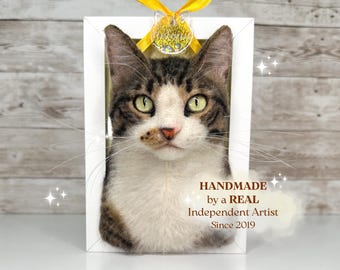 May include: A needle-felted cat portrait in a white rectangular frame with a yellow ribbon at the top. The cat has brown tabby markings, a white chest and face, and green eyes. Text on the image reads: HANDMADE by a REAL Independent Artist Since 2019.