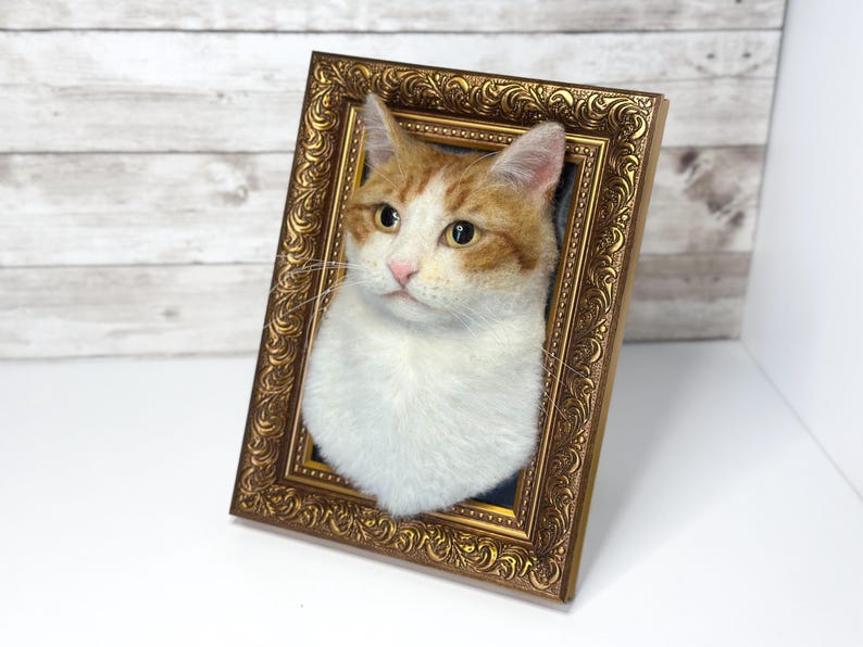 May include: A three-dimensional needle felted cat portrait featuring a white and orange tabby cat head. The cat has amber eyes and a pink nose, set within an ornate, rectangular, metallic gold-colored picture frame with intricate scrollwork.