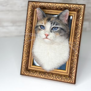 May include: A three-dimensional cat portrait sculpture featuring a calico cat with blue eyes, a pink nose, and white chest fur, mounted inside an ornate, rectangular, antique-style gold-toned frame with intricate embossed scrollwork patterns.