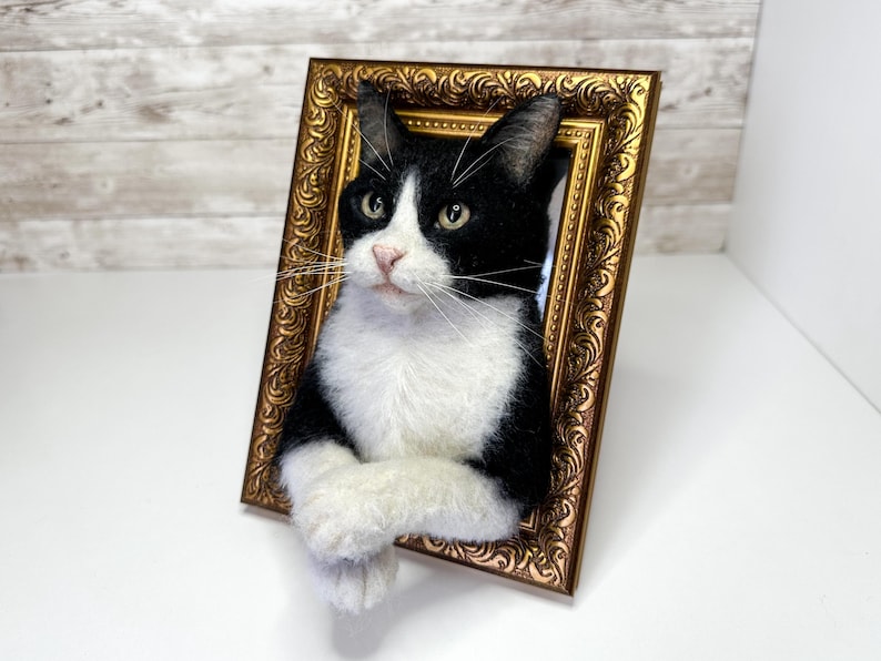 Una escultura de gato de lana fieltrada en blanco y negro que emerge de un marco de fotos dorado y ornamentado. El gato tiene el pecho blanco, patas blancas cruzadas sobre el borde inferior del marco y una cabeza negra con marcas blancas.