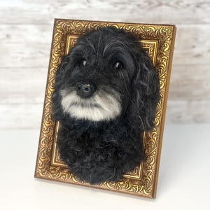 May include: A three-dimensional needle felted sculpture of a black dog with a white muzzle, mounted inside an ornate, rectangular gold-coloured frame. The dog has curly black fur, dark eyes, and a black nose, creating a realistic portrait effect.