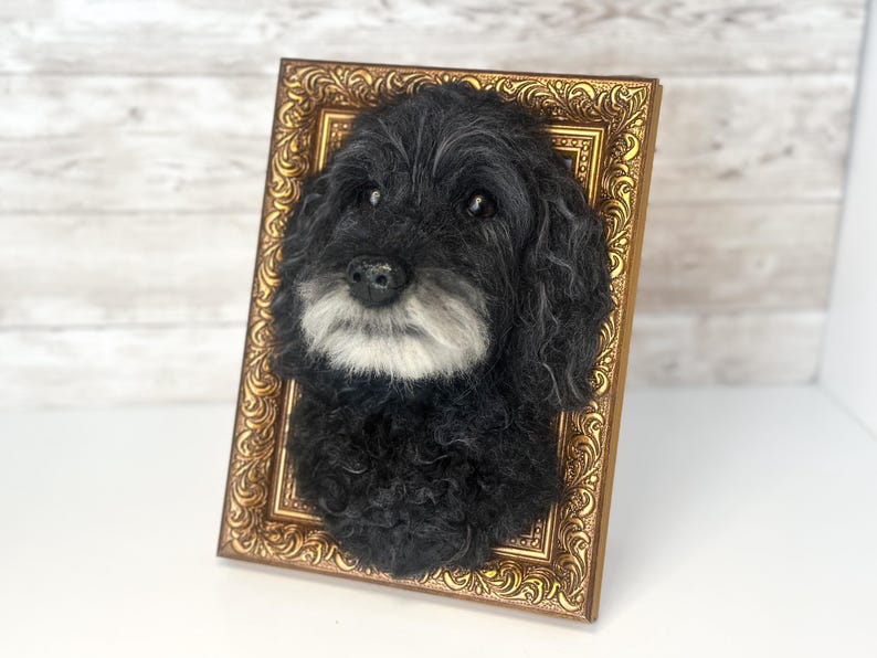 May include: A three-dimensional needle felted sculpture of a black dog with a white muzzle, mounted inside an ornate, rectangular gold-coloured frame. The dog has curly black fur, dark eyes, and a black nose, creating a realistic portrait effect.