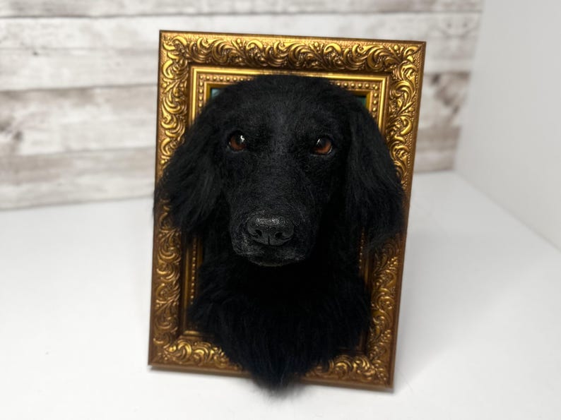 May include: A three-dimensional wall sculpture of a black dog head with long, soft fur and brown eyes, mounted inside a rectangular, ornate, metallic gold-coloured frame with intricate embossed scrollwork patterns along the border.