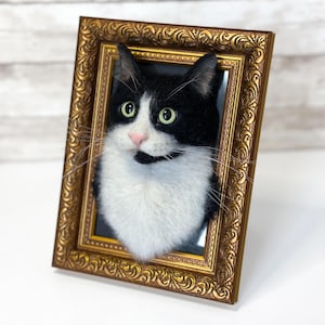 May include: A needle-felted black and white cat portrait mounted inside an ornate, rectangular gold-colored frame. The cat has green eyes, a pink nose, and long white whiskers, with its head and chest protruding from the frame's center.