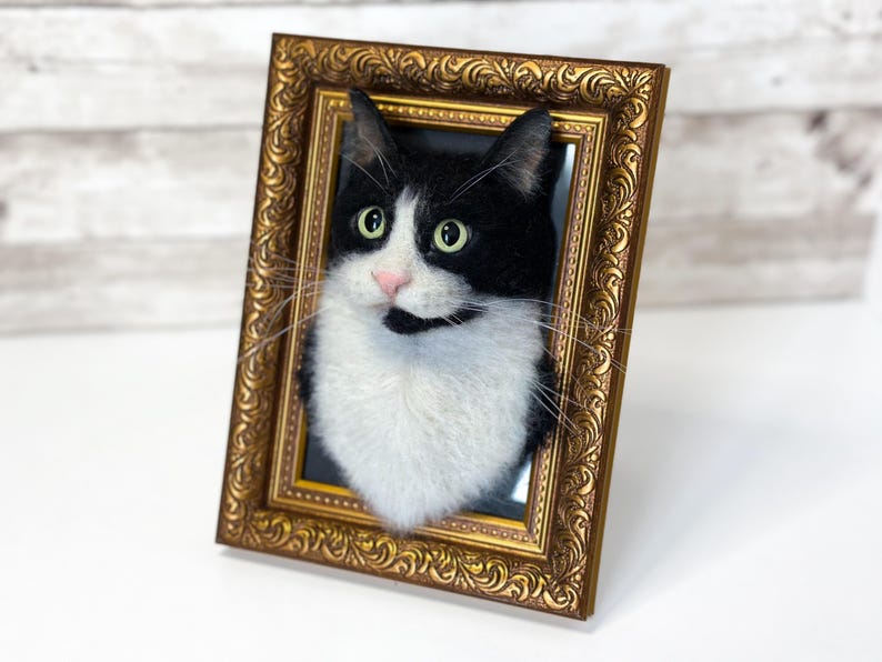 May include: A needle-felted black and white cat portrait mounted inside an ornate, rectangular gold-colored frame. The cat has green eyes, a pink nose, and long white whiskers, with its head and chest protruding from the frame's center.