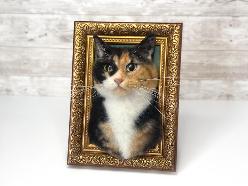 May include: A three-dimensional needle felted calico cat portrait mounted inside an ornate, rectangular gold-toned frame. The cat features a mix of black, orange, and white fur with long, thin whiskers, set against a solid teal background.