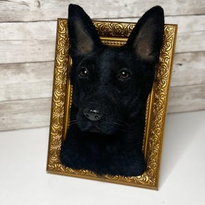 May include: A three-dimensional needle felted sculpture of a black dog head mounted inside an ornate, rectangular gold-coloured frame. The dog has pointed ears, dark eyes, and a textured black coat, creating a realistic portrait effect.