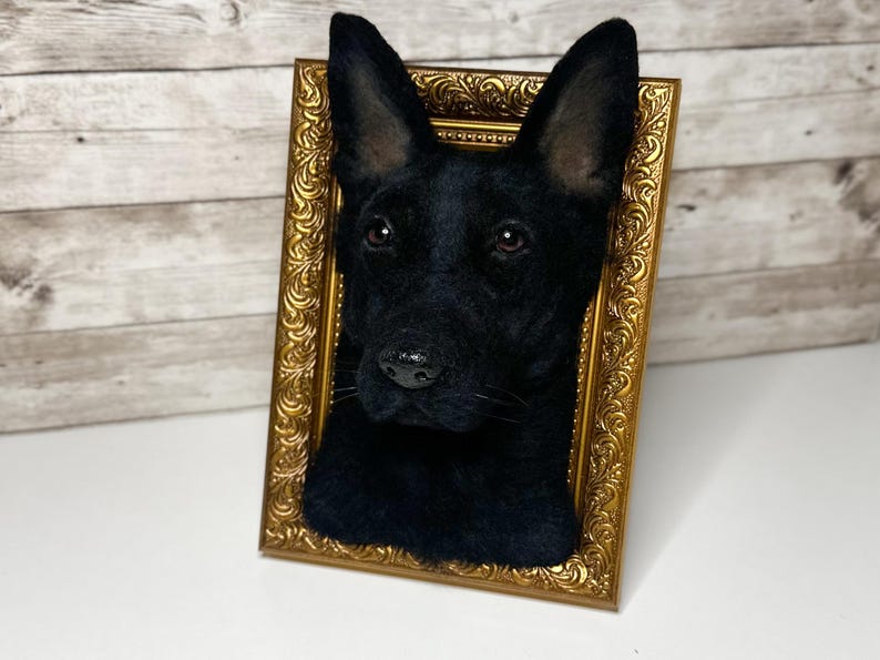 May include: A three-dimensional needle felted sculpture of a black dog head mounted inside an ornate, rectangular gold-coloured frame. The dog has pointed ears, dark eyes, and a textured black coat, creating a realistic portrait effect.