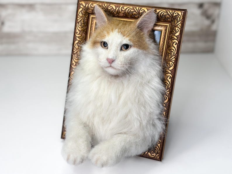 May include: A needle-felted sculpture of a long-haired cat with white and ginger fur, positioned within an ornate, rectangular, metallic bronze-colored picture frame. The cat has a white face, ginger ears, and white paws resting on the frame edge.