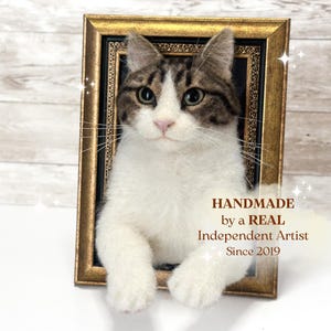 Pode incluir: Uma escultura de gato em feltro com pelo branco e tigrado castanho, posicionada dentro de uma moldura retangular dourada e preta. O texto HANDMADE by a REAL Independent Artist Since 2019 está impresso em castanho no canto inferior direito.