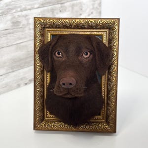 May include: A three-dimensional needle felted sculpture of a chocolate Labrador retriever head mounted inside an ornate, rectangular gold-coloured frame. The dog has dark brown fur, light brown eyes, and a textured appearance, creating a realistic portrait.