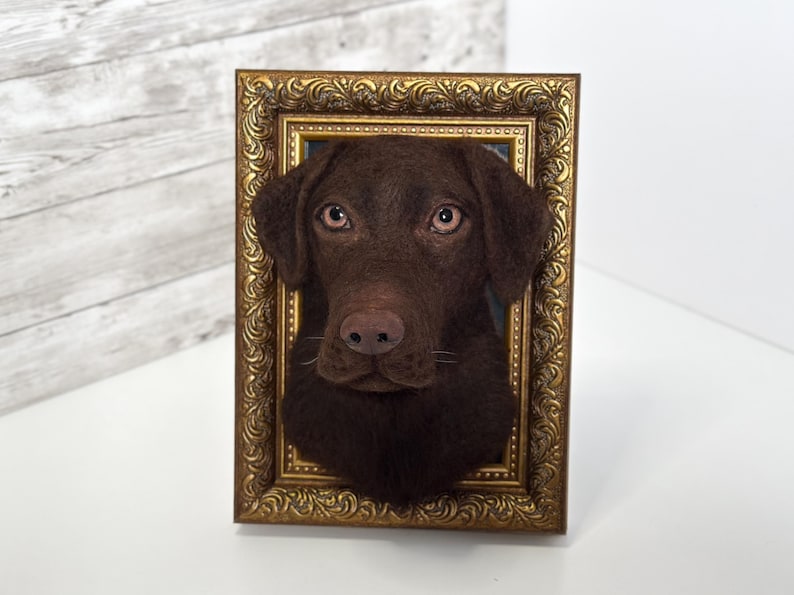 May include: A three-dimensional needle felted sculpture of a chocolate Labrador retriever head mounted inside an ornate, rectangular gold-coloured frame. The dog has dark brown fur, light brown eyes, and a textured appearance, creating a realistic portrait.