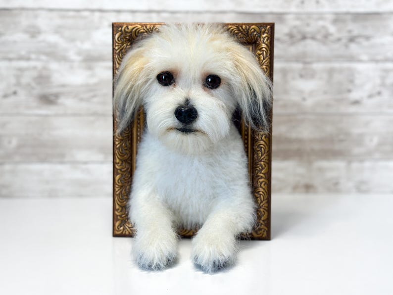 May include: A plush dog figurine with white fur and dark-tipped ears, positioned inside a rectangular, ornate, metallic bronze-coloured picture frame. The dog has dark, round eyes and a black nose, with its front paws resting at the bottom edge.