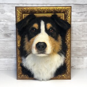 May include: A needle-felted dog head sculpture featuring black, white, and tan fur, mounted within an ornate, rectangular gold-coloured frame. The dog has brown eyes, a black nose, and a white blaze extending from the forehead to the chest.