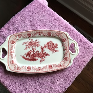 May include: An off-white rectangular serving platter with a red toile design depicting a rural scene. The platter has decorative handles and a red floral border. It rests on a purple towel. The platter is approximately 30 cm long.