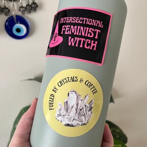 May include: A light green water bottle with two stickers. The top sticker reads "INTERSECTIONAL FEMINIST WITCH" in pink text on a black background. The bottom sticker says "FUELED BY CRYSTALS & COFFEE" with a crystal illustration.