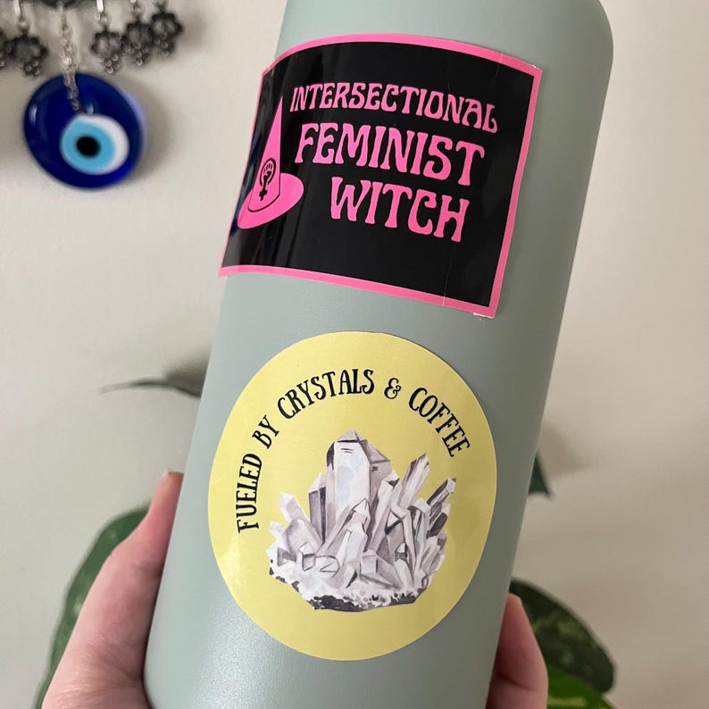May include: A light green water bottle with two stickers. The top sticker reads "INTERSECTIONAL FEMINIST WITCH" in pink text on a black background. The bottom sticker says "FUELED BY CRYSTALS & COFFEE" with a crystal illustration.