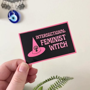 May include: A black sticker with a pink border and text that reads "INTERSECTIONAL FEMINIST WITCH." The sticker features a pink witch hat with a feminist symbol. The sticker is held by a hand.