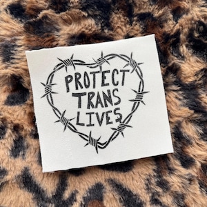 May include: A square, off-white patch with the words "PROTECT TRANS LIVES" in bold, black letters, encircled by a heart-shaped barbed wire design. The patch rests on a brown and black leopard print background.