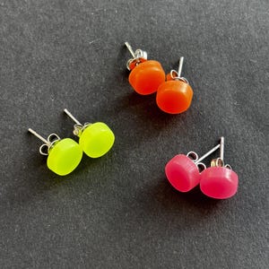 May include: Three pairs of stud earrings in neon yellow, bright orange, and hot pink. Each pair has a rounded square shape with a silver post. The earrings are displayed on a dark grey surface.
